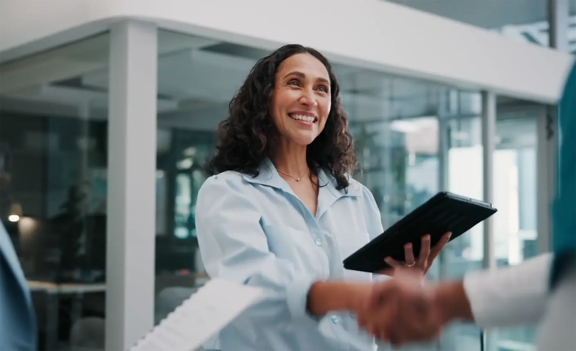 Smiling woman holding a tablet while shaking hands with someone in a modern office, representing a business agreement or professional partnership.