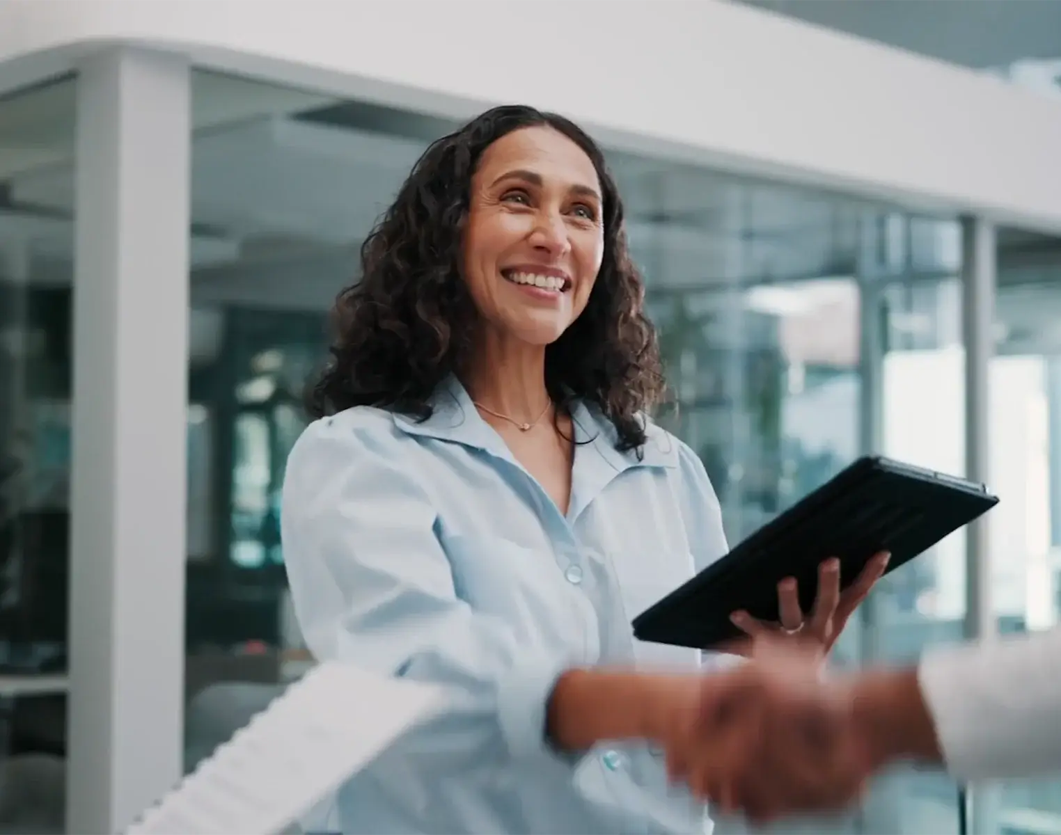 Smiling woman holding a tablet while shaking hands with someone in a modern office, representing a business agreement or professional partnership.
