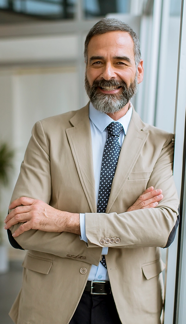 Smiling middle-aged man with a beard, wearing a beige suit and tie, standing with arms crossed by a window in a bright office setting.