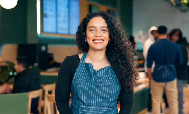 Smiling woman wearing an apron standing in a café or restaurant, with coworkers and customers in the background, representing hospitality or food service work.”