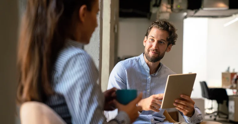 Man holding a tablet and talking with a woman holding a cup, engaged in a casual discussion in an office or collaborative workspace.