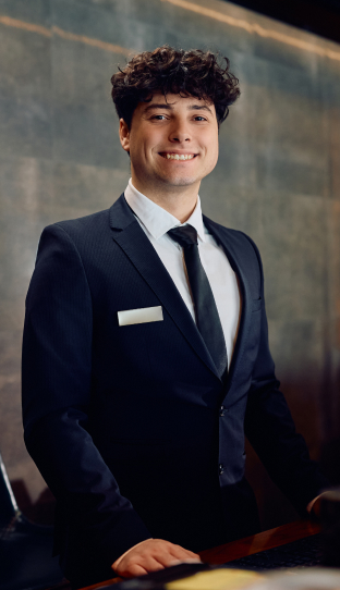 Smiling man in a suit and tie standing at a reception desk, likely a hotel or front desk employee, representing customer service or hospitality.