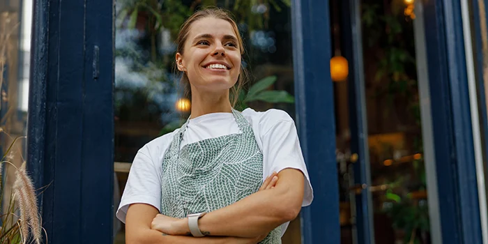 Smiling café worker wearing an apron standing at the entrance of a shop with arms crossed and greenery in the background.