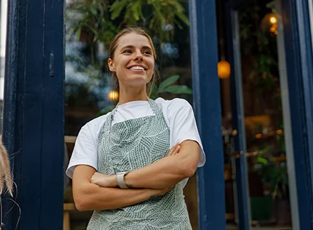 Smiling café worker wearing an apron standing at the entrance of a shop with arms crossed and greenery in the background.