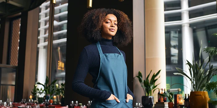 A confident woman with curly hair stands in a restaurant wearing a dark top and a teal apron, hands in pockets, with tables and warm lighting in the background.