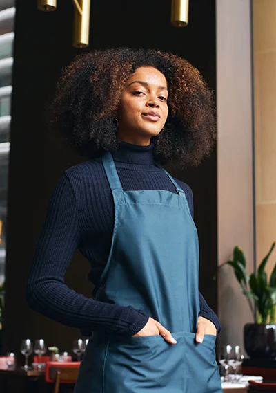 A confident woman with curly hair stands in a restaurant wearing a dark top and a teal apron, hands in pockets, with tables and warm lighting in the background.