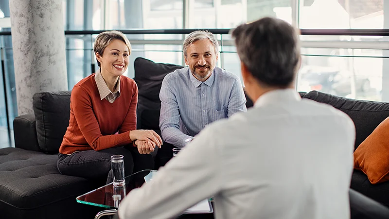 A smiling couple sits on a couch in a modern office, talking with a professional seated across from them, suggesting a consultation or meeting.