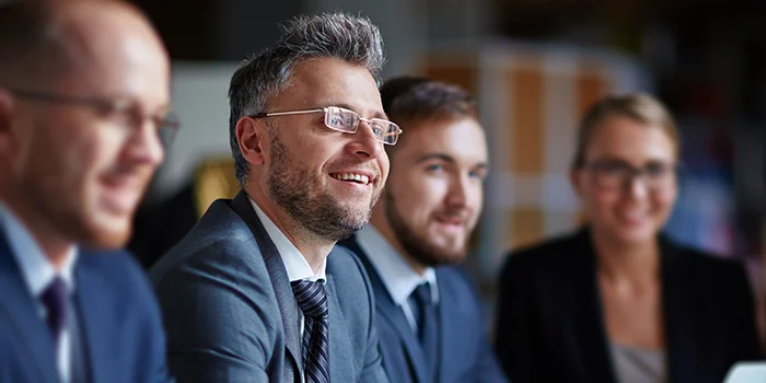 Group of business professionals in suits sitting at a table, smiling and engaged in a meeting, with a laptop and documents in front of them.