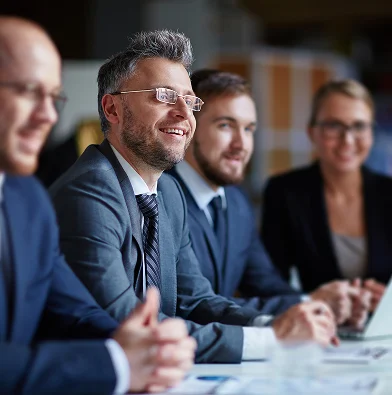 Group of business professionals in suits sitting at a table, smiling and engaged in a meeting, with a laptop and documents in front of them.
