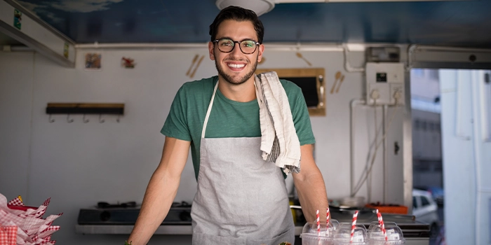 Smiling food vendor wearing an apron and glasses standing behind a counter in a small kitchen or food stall.