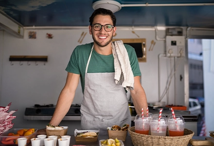 Smiling food vendor wearing an apron and glasses standing behind a counter in a small kitchen or food stall.