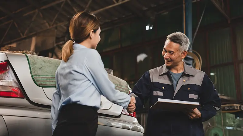 Mechanic shaking hands with a customer next to a car in a garage, holding a clipboard as they finalize service details.