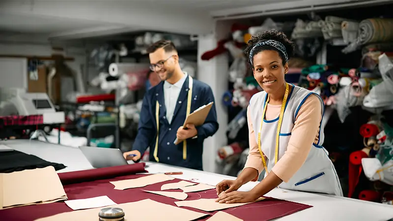 Two garment workers in a textile workshop cutting fabric patterns on a table, with rolls of fabric and tools in the background.
