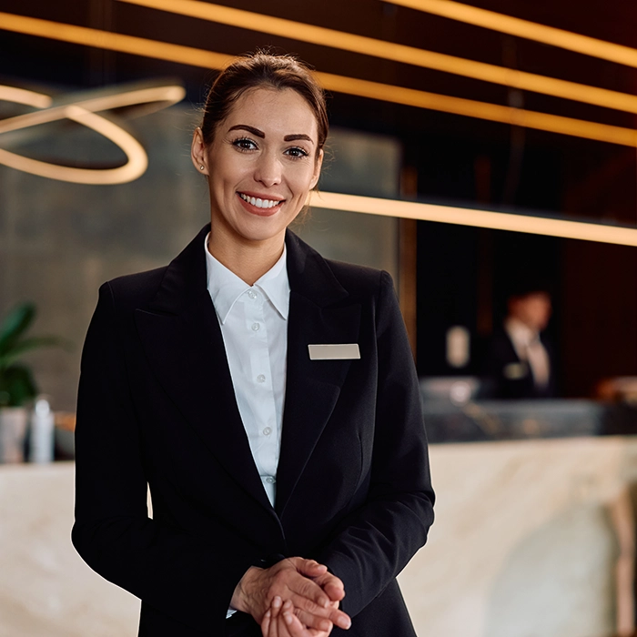 Smiling hotel receptionist in a formal uniform standing at a front desk in a modern lobby.