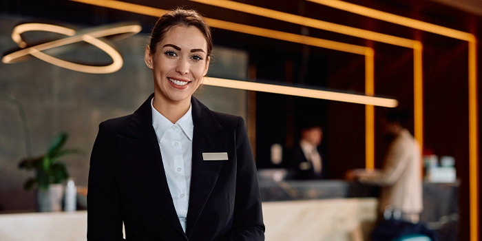 Smiling hotel receptionist in a formal uniform standing at a front desk in a modern lobby.