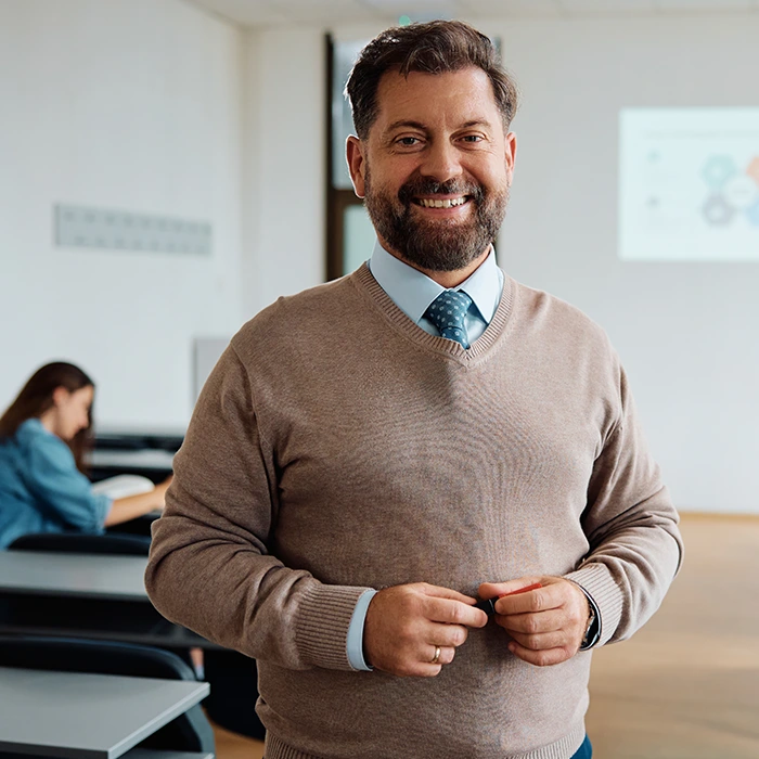 Smiling teacher standing in a classroom holding a marker, with students seated in the background.