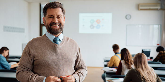 Smiling teacher standing in a classroom holding a marker, with students seated in the background.