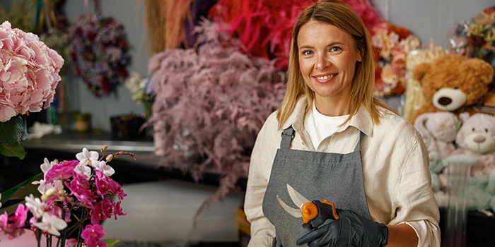 Florist wearing an apron and gloves holding pruning shears in a flower shop, surrounded by colorful floral arrangements and decorations.