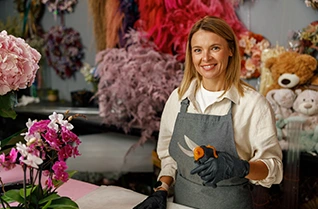 Florist wearing an apron and gloves holding pruning shears in a flower shop, surrounded by colorful floral arrangements and decorations.