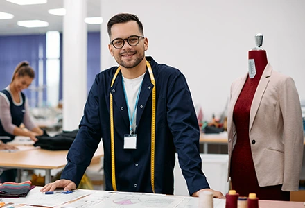 Fashion designer with a measuring tape around his neck standing in a studio next to a mannequin dressed in a blazer, with a colleague working in the background.