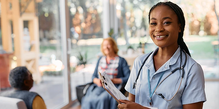 Smiling nurse holding a clipboard in a clinic waiting area, with patients seated in the background.