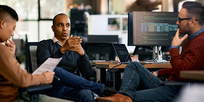 Three colleagues in an office discussing a project, with one speaking while others listen, and a computer screen displaying code in the background.
