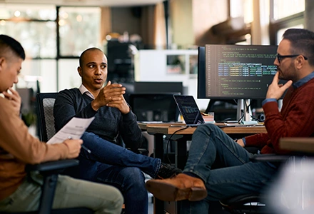 Three colleagues in an office discussing a project, with one speaking while others listen, and a computer screen displaying code in the background.