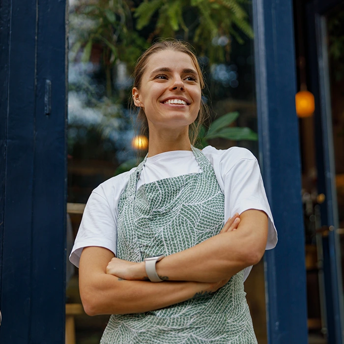 Smiling café worker wearing an apron standing at the entrance of a shop with arms crossed and greenery in the background.