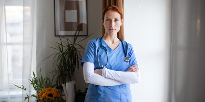 Healthcare professional in blue scrubs with a stethoscope standing with arms crossed in a clinic setting.