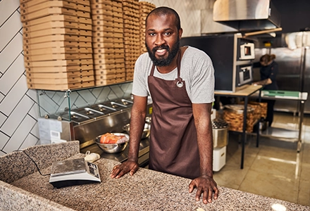 Restaurant worker wearing an apron preparing food in a commercial kitchen, with stacked pizza boxes and equipment in the background.