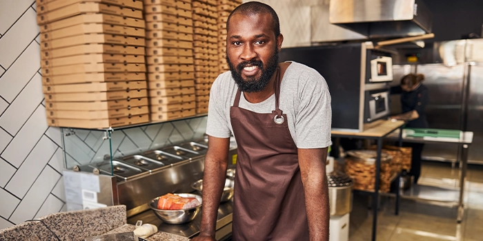 Restaurant worker wearing an apron preparing food in a commercial kitchen, with stacked pizza boxes and equipment in the background.