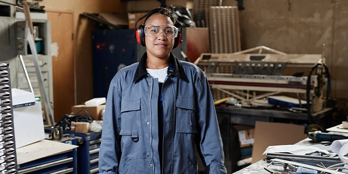 Factory worker wearing safety glasses and ear protection standing in an industrial workshop with machinery and tools in the background.