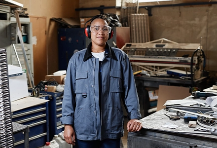 Factory worker wearing safety glasses and ear protection standing in an industrial workshop with machinery and tools in the background.