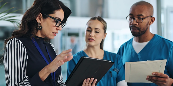 Three healthcare professionals reviewing information together, with one woman holding a tablet and two colleagues in scrubs looking on, one holding a document.