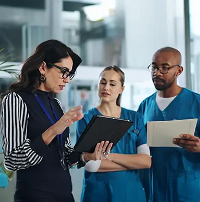 Three healthcare professionals reviewing information together, with one woman holding a tablet and two colleagues in scrubs looking on, one holding a document.
