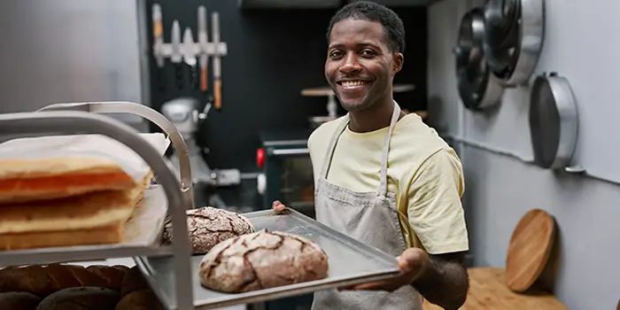 Smiling baker holding a tray of freshly baked bread in a kitchen, with loaves and baking tools visible in the background.
