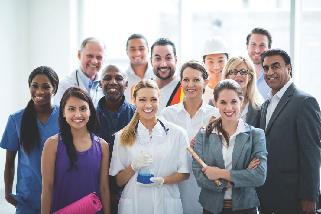 Diverse group of professionals smiling at the camera, including a doctor, construction worker, scientist holding a flask, businesspeople, and fitness enthusiast, standing together in a bright indoor setting.
