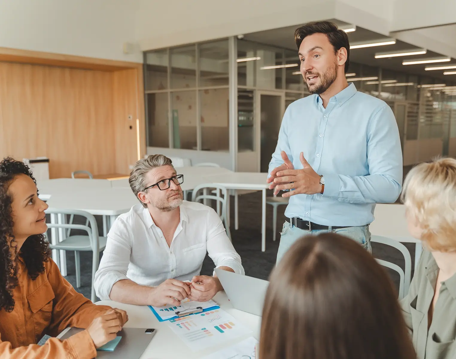 A man standing and speaking to a small group of colleagues during a meeting in a modern office, gesturing with his hands while others sit around a table with documents and a laptop, listening attentively.