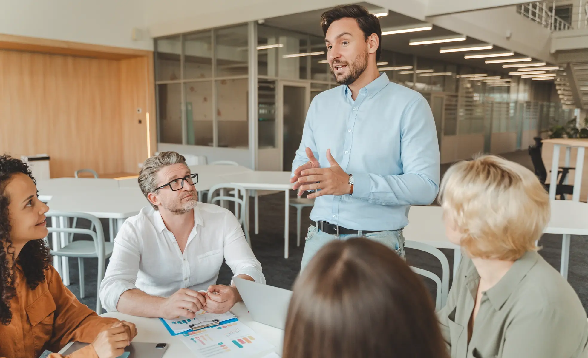 A man standing and speaking to a small group of colleagues during a meeting in a modern office, gesturing with his hands while others sit around a table with documents and a laptop, listening attentively.